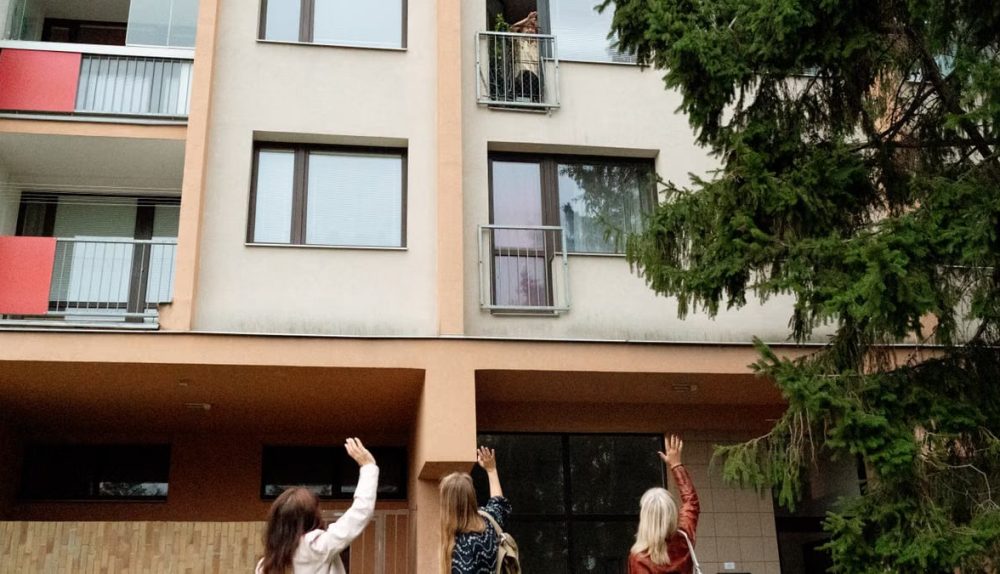 Three women waving to an elderly woman on an apartment balcony in Prague.